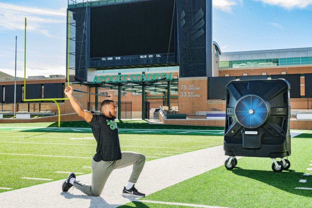 A man kneels triumphantly on a football field with an evaporative cooling system. A stadium screen and goalposts are in the background.