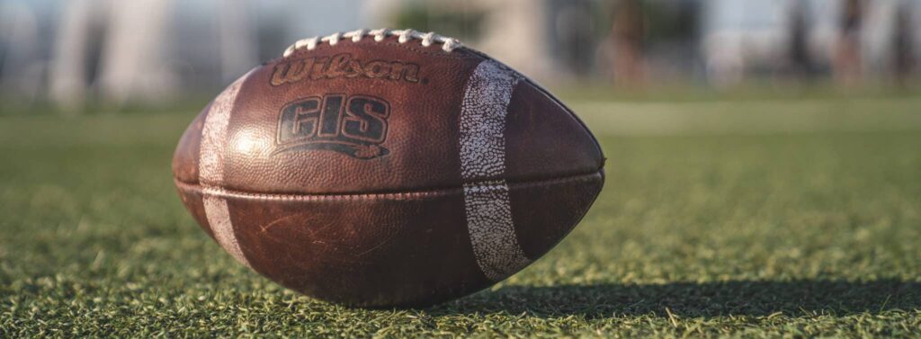 A weathered Wilson GLS football lies on grass, suggesting past play; blurred background hints at an outdoor sideline setting.