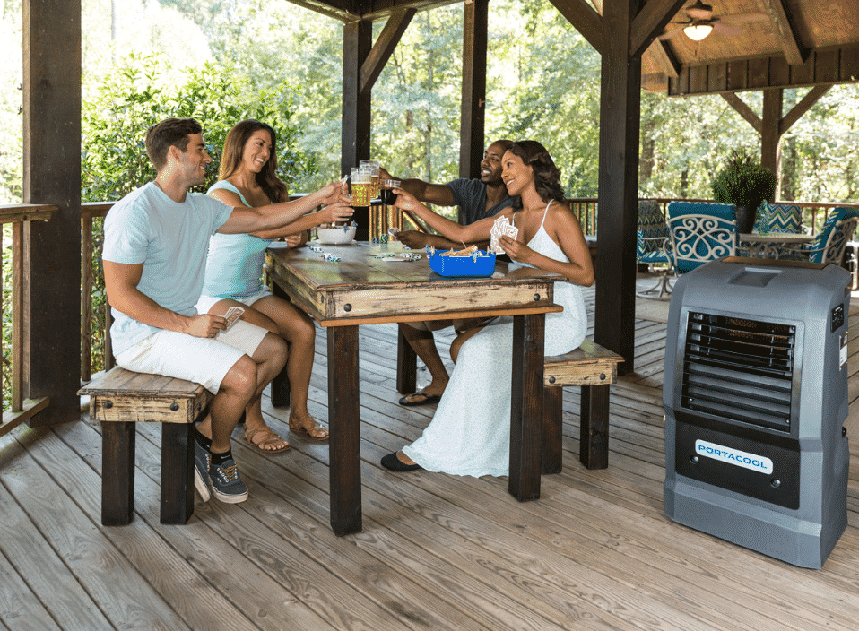 Four people on a porch toast the start of summer. Nearby, a portable evaporative cooler ensures comfort in the tree-filled setting.