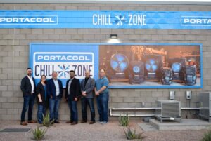 A group of seven people in business casual attire stands before a "Portacool Chill Zone" sign and a large banner. Lively scene with vibrant plants.