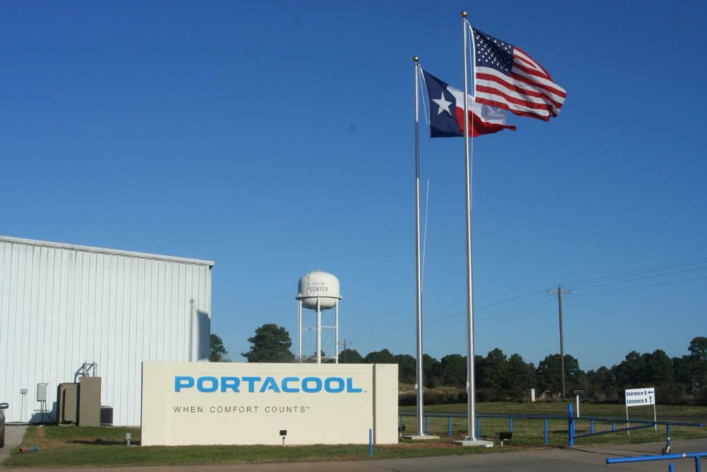 Sign at industrial entrance reads "Portacool When Comfort Counts." U.S. and Texas flags on flagpoles, with trees and water tower in the background.