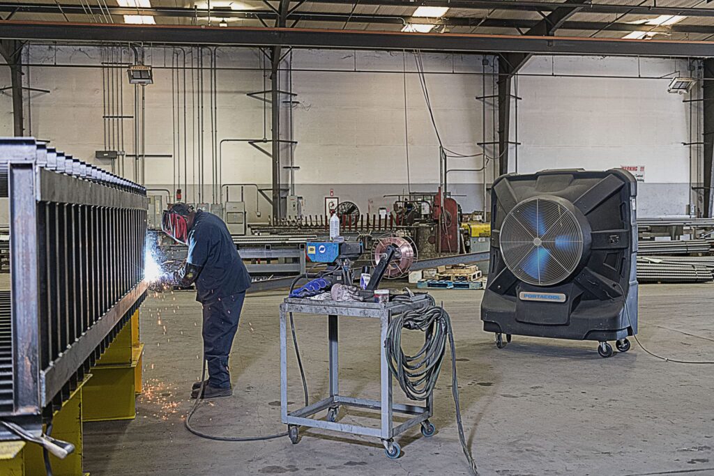 A welder in protective gear works with sparks flying. Tools and cables on a cart nearby. Shelves and equipment emphasize safety in preventing heat stress.