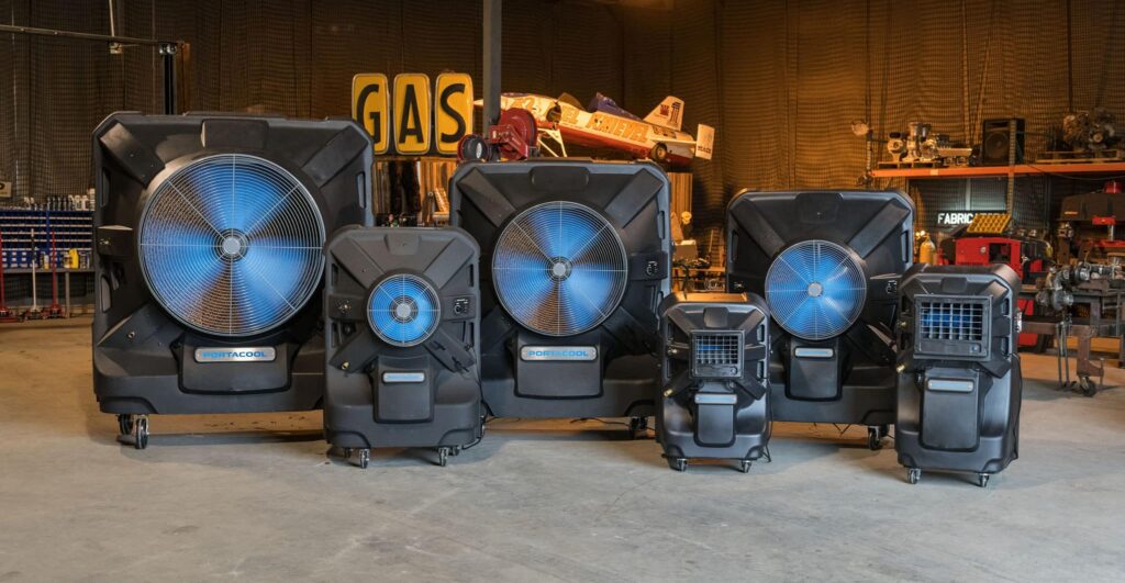 Five industrial units with large blue blades lineup in a workshop, demonstrating evaporative cooling. Tools and a "GAS" sign are in the background.