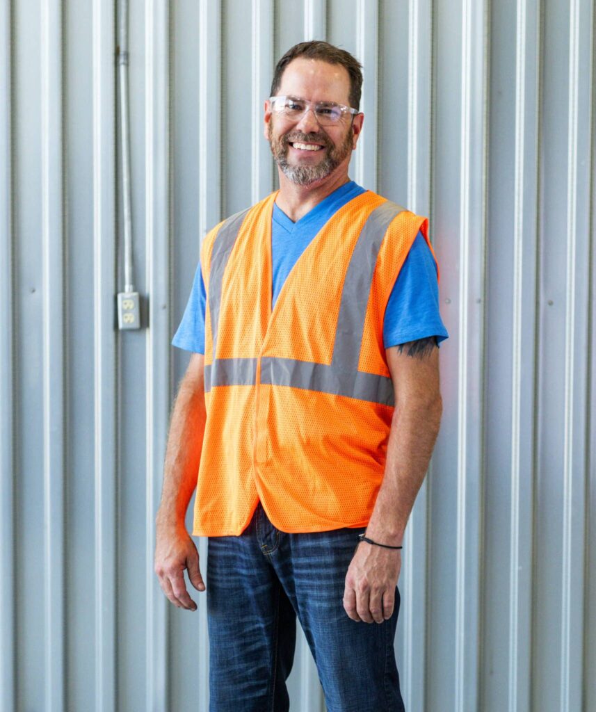 A man in safety glasses, blue shirt, and orange safety vest smiles in front of corrugated metal siding, highlighting hazard recognition.