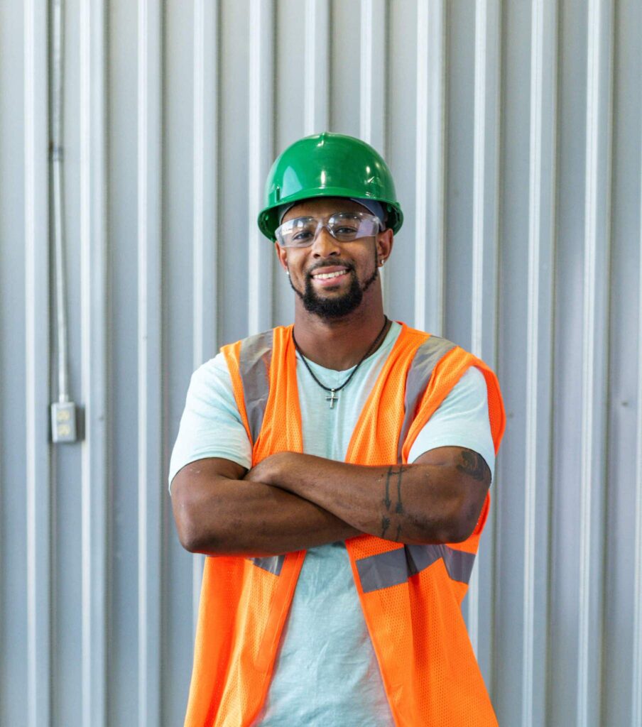 A confident person wearing a green hard hat, safety glasses, and an orange vest stands against vertical metal panels.