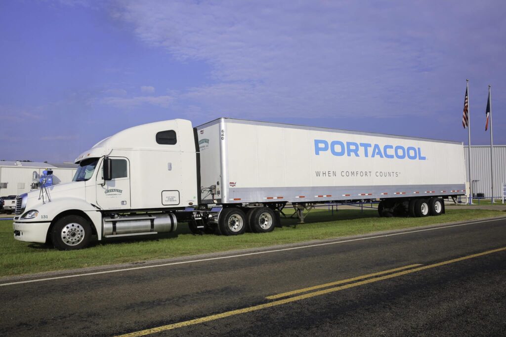 White semi-truck with "Portacool" branding parked on grass near a road; two flags in the background under a clear sky.