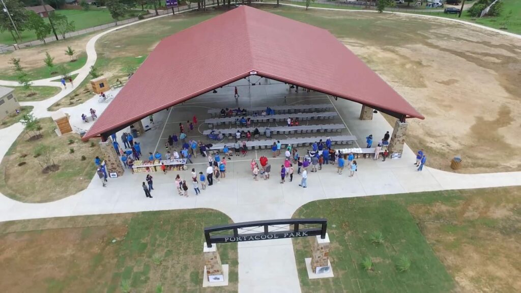 Aerial view of a pavilion with a red roof in Portacool Park. People celebrate an anniversary under the open-air structure, surrounded by grass and pathways.
