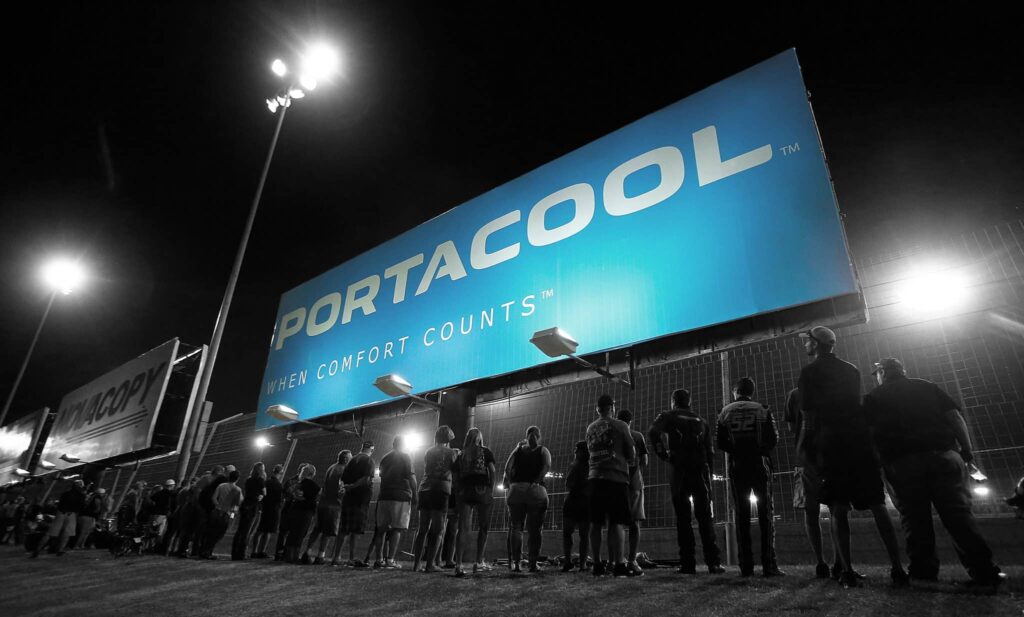A group stands before an illuminated billboard at night with "PORTACOOL" in bold letters and the tagline "WHEN COMFORT COUNTS," evoking a motorsport start.