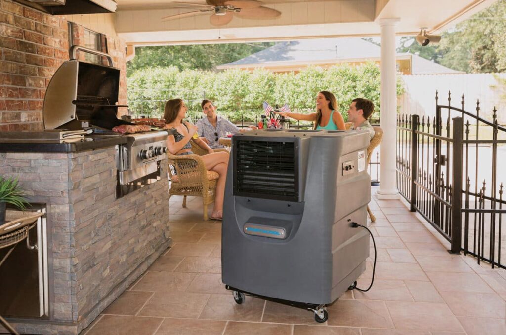 A group of four relaxes on a covered patio near a stone outdoor kitchen with a grill, surrounded by lush trees, creating the perfect Portacool Porch retreat.