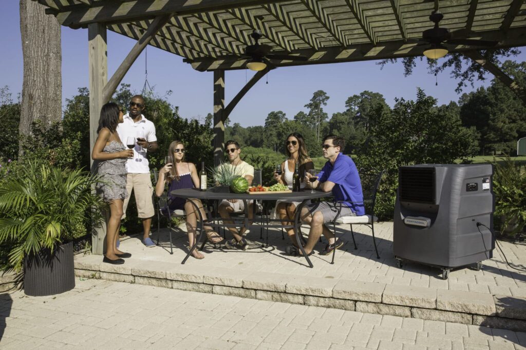 Six people chat and enjoy drinks under a pergola surrounded by lush greenery on a sunny day.