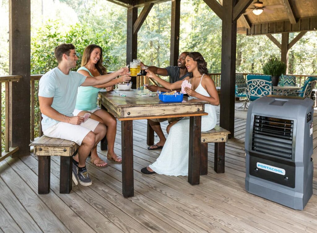 Four people toast with drinks at a table on a covered deck. A Portacool unit ensures comfort in their wooded summer gathering.