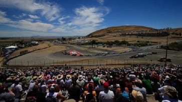 Spectators at Sonoma Raceway watch a car race on a winding track, seated in bleachers under a clear blue sky with a large hill in the background.