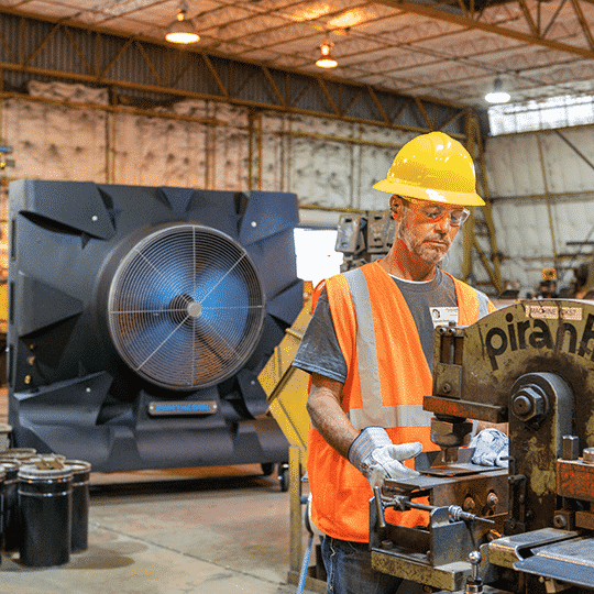 A worker in a hard hat and safety vest operates a metal cutting machine in a well-lit industrial workshop with large equipment.