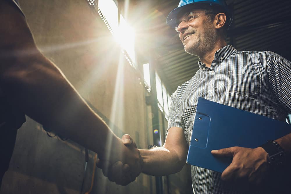 In sunlight, a man in a hard hat and glasses holds a blue clipboard while shaking hands with another person, symbolizing a successful partnership.