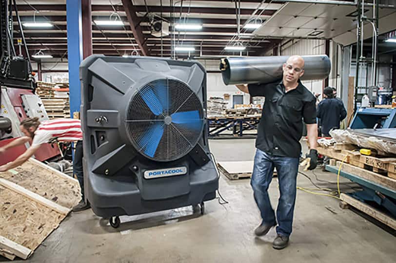 A man carries rolled material in a warehouse with pallets, machinery, and a large Portacool unit.