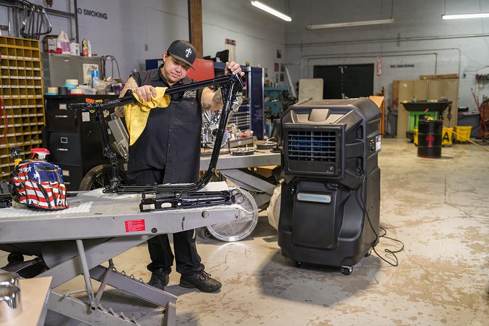 A person in a workshop wipes a bicycle frame with a yellow cloth. Nearby, tools are scattered and there's a Portacool air cooler.