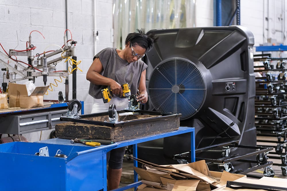 In a busy factory, a person in safety glasses uses a drill on wood. Tools and equipment are scattered on blue tables, creating an industrious vibe.