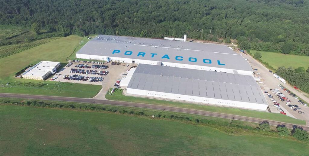 Aerial view of an industrial building with "Portacool" on the roof, surrounded by parking lots, roads, green fields, and distant forests.