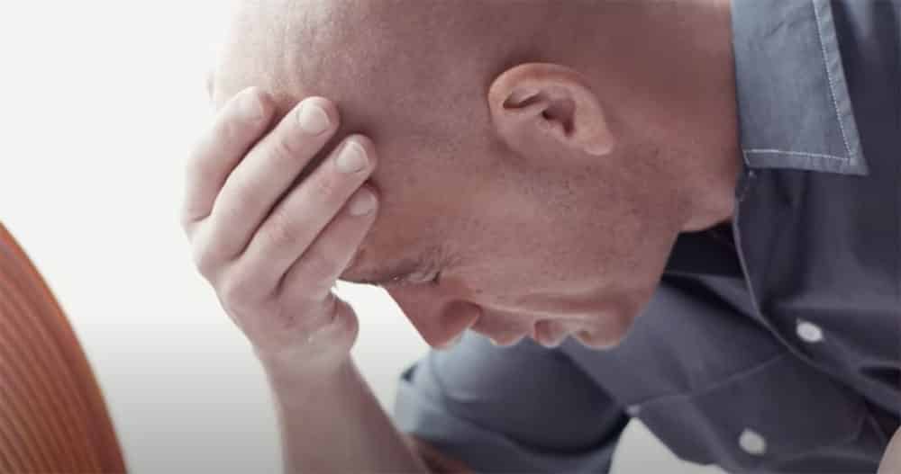 A bald man with a beard, in a gray shirt, sits thoughtfully with his hand on his forehead against a blurred background.
