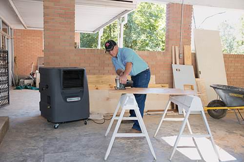 A person in a cap focuses on woodworking a large plank on white sawhorses, surrounded by tools in an OSHA-compliant outdoor area.