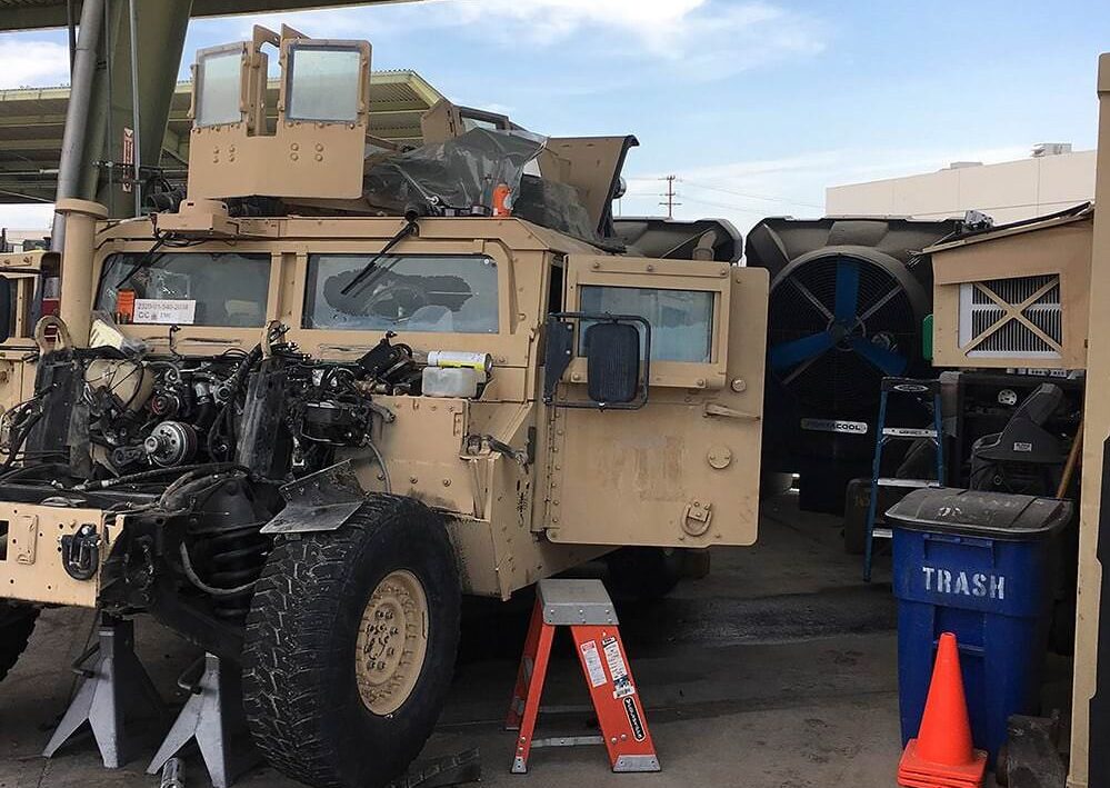A tan military Humvee under a shelter with its front engine disassembled, a step ladder, and a blue trash bin in the background.