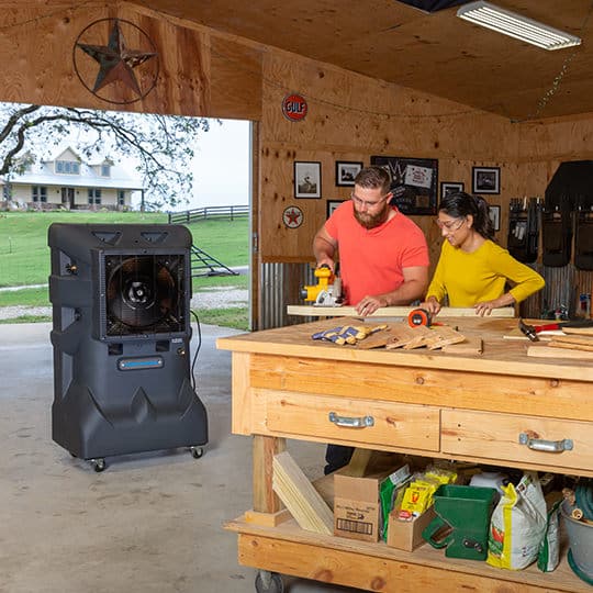 A man and woman work in a wooden shed; he uses a tool on a plank with her assistance. A large house is visible nearby, inspiring their organized workspace.