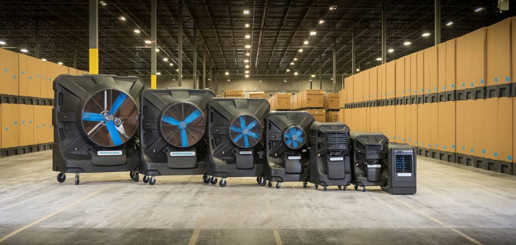 Large black industrial cooling units with blue blades on wheels in a warehouse, surrounded by stacked cardboard boxes.