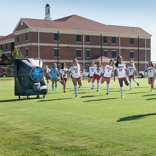 A women's soccer team in red and white jogs energetically on a grassy field, with a brick building and clear blue sky in the background.