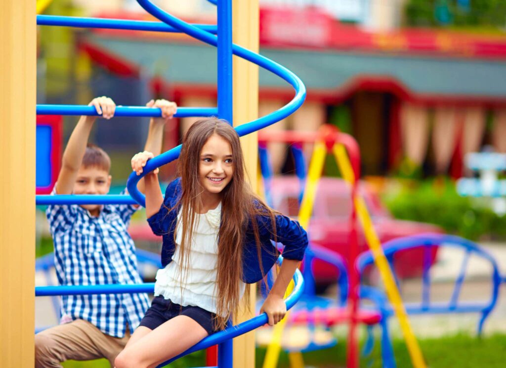 Children play on a sunny day; girl climbs a spiral structure, boy in blue checkered shirt holds onto playground equipment.