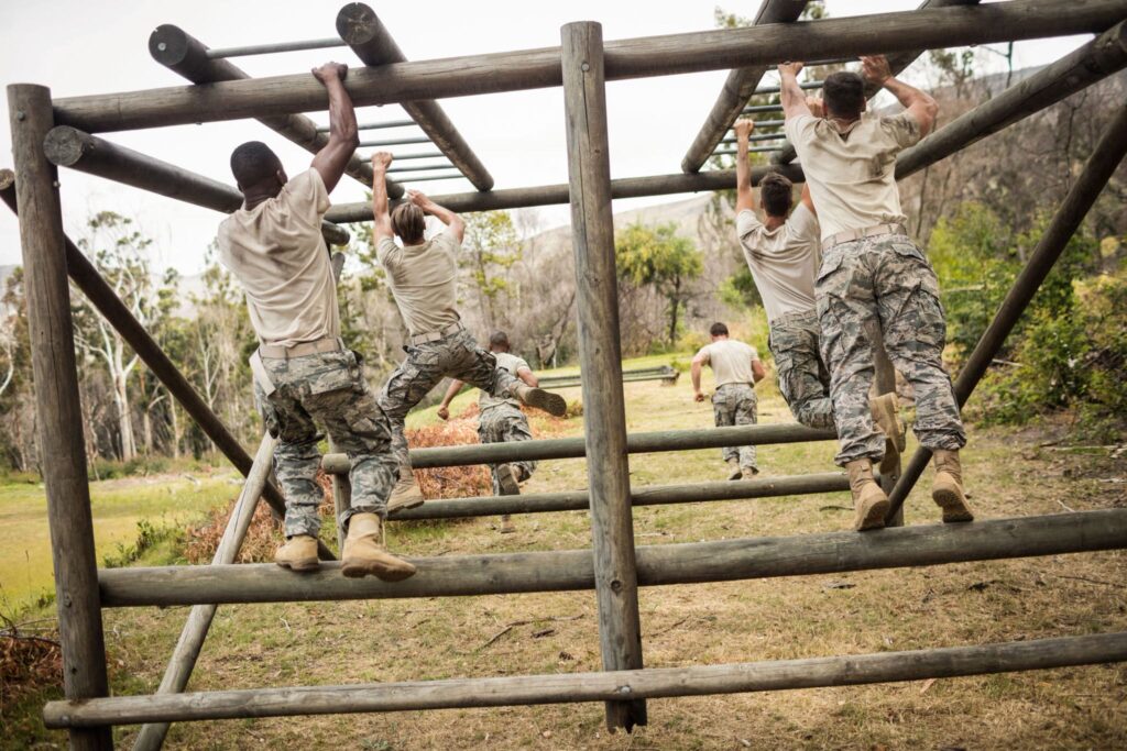 U.S. troops in military attire tackle an outdoor monkey bar obstacle course, wearing camouflage pants and boots with trees in the background.