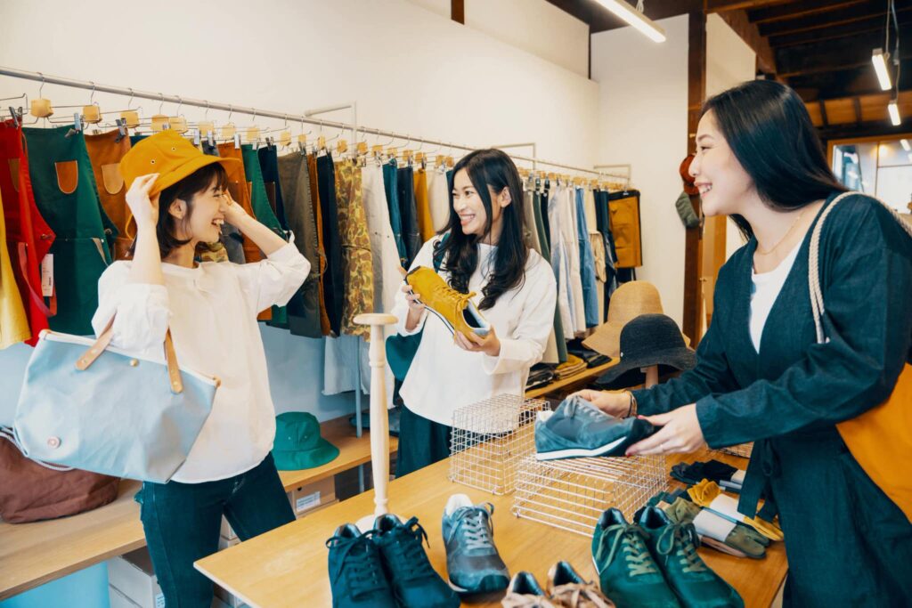 Three women shop in a clothing store. One tries on a yellow hat for hot weather, another holds shoes, and the third examines sneakers with clothes in the background.