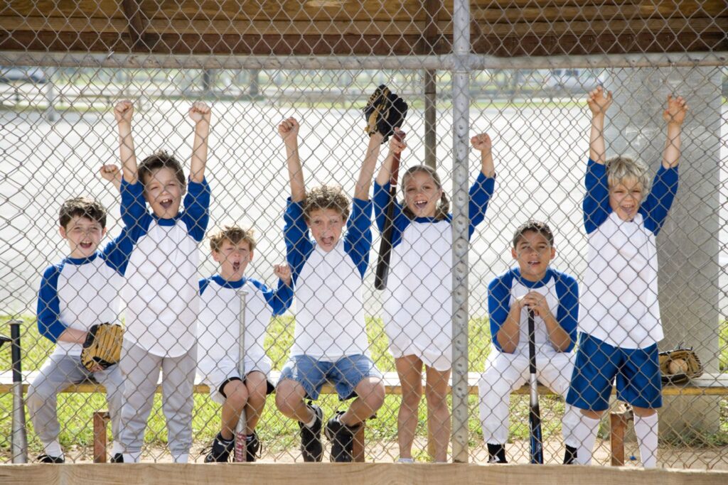 Seven uniformed kids cheerfully raise arms on a bench behind a chain-link fence, awaiting snacks at the baseball game.