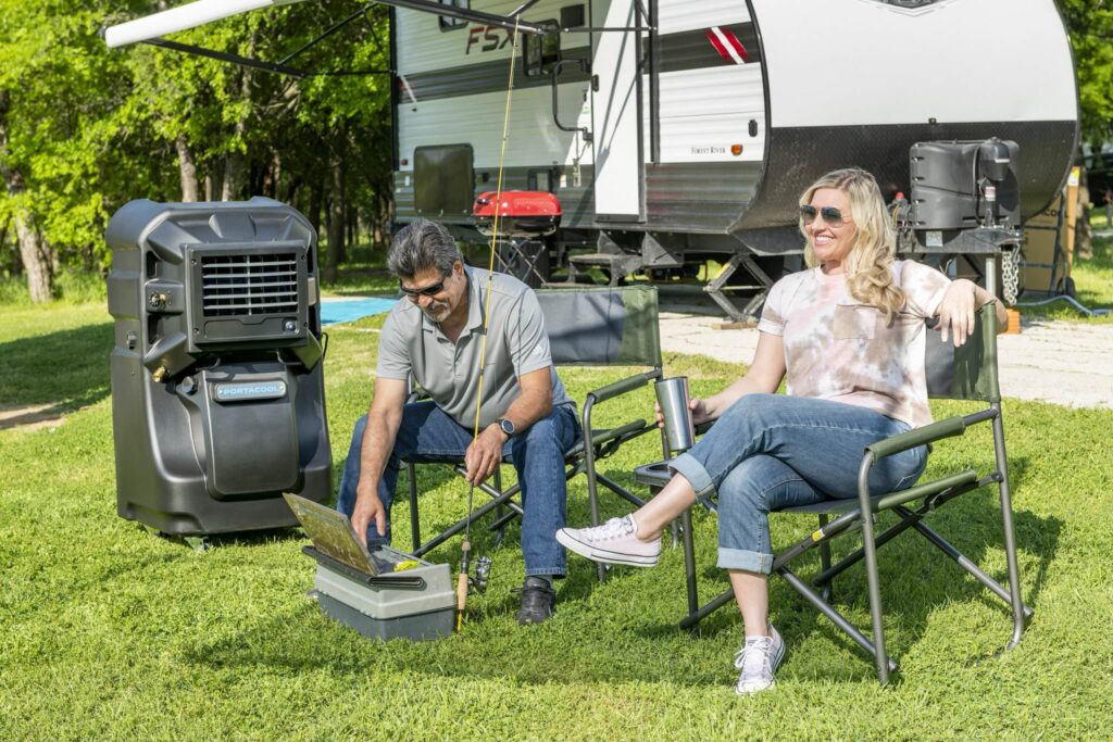 A couple enjoys relaxing by their RV. He adjusts a Portacool; she holds a tumbler. Tall trees sway in the background, enhancing the tranquility.