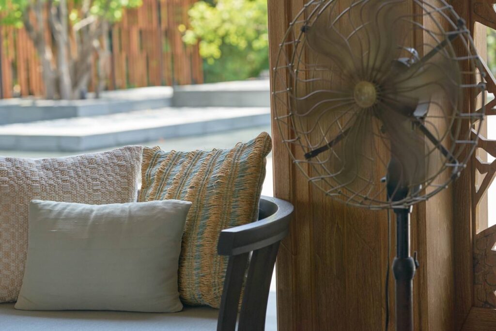 Outdoor seating with two pillows on a wooden bench. A vintage metal fan stands nearby, with greenery and steps creating a serene backdrop.