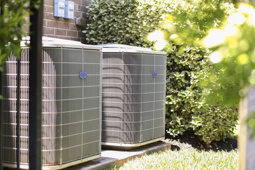 Two HVAC units on concrete beside a brick wall, surrounded by green foliage, showcasing the answer to does air conditioning use gas.