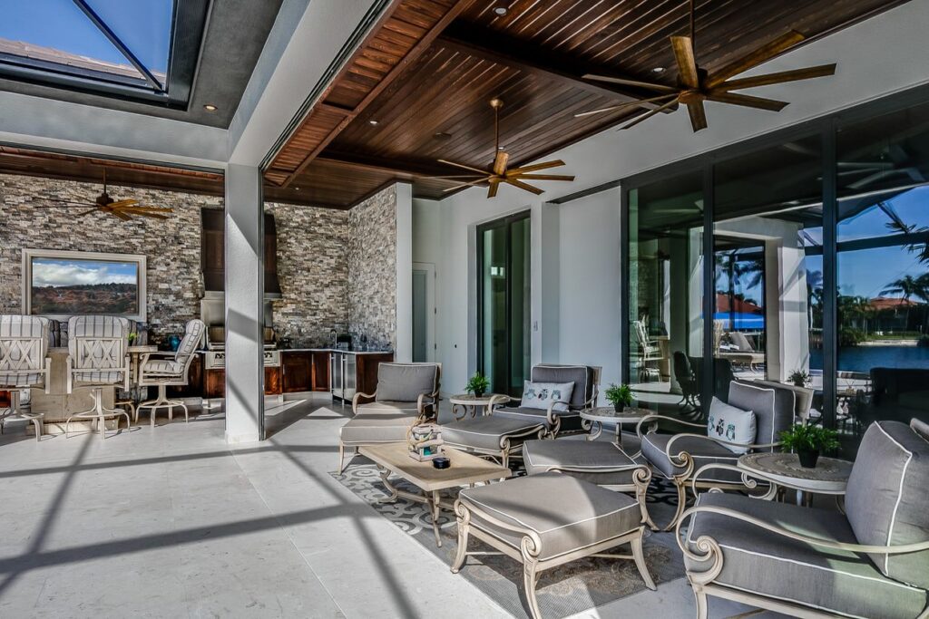 Outdoor patio with cushioned seating, coffee tables, and stone accent wall. Glass doors reveal pool and palm trees; wooden ceiling with efficient Portacools.
