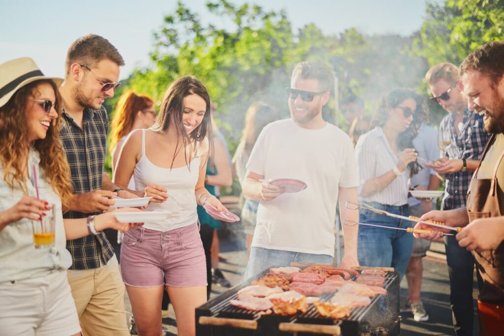 A group enjoys a sunny barbecue on the patio, smiling with plates while meat grills. Misting systems cool the scene, greenery in the background. which is best for my patio?