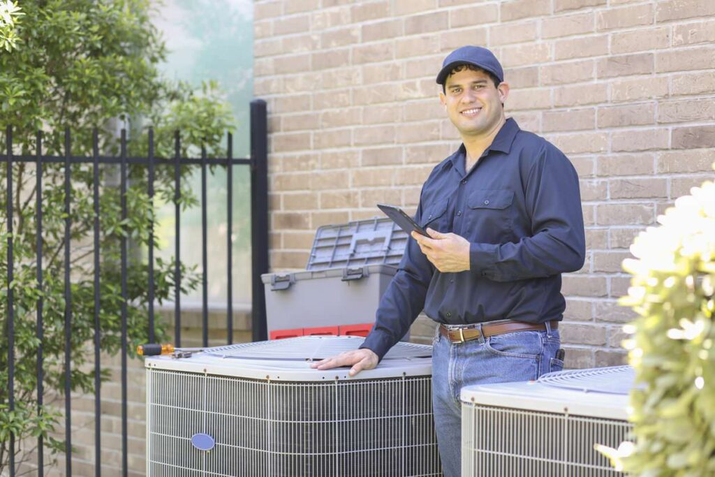 A smiling technician in blue holds a tablet next to two large outdoor units, with a brick wall and greenery in the background.