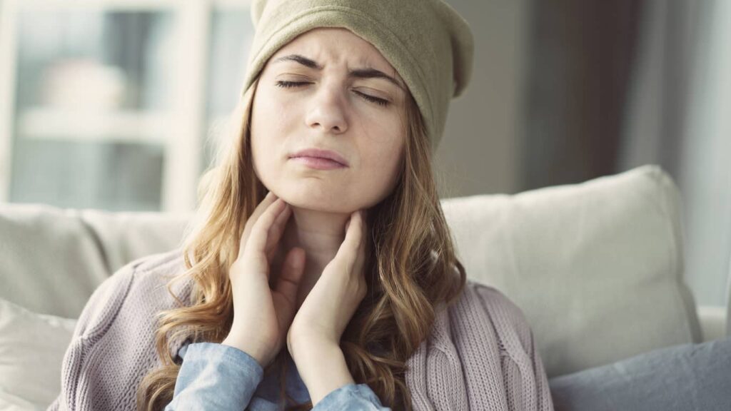 Woman in green hat and purple sweater touches her throat wondering can air conditioning cause a sore throat.