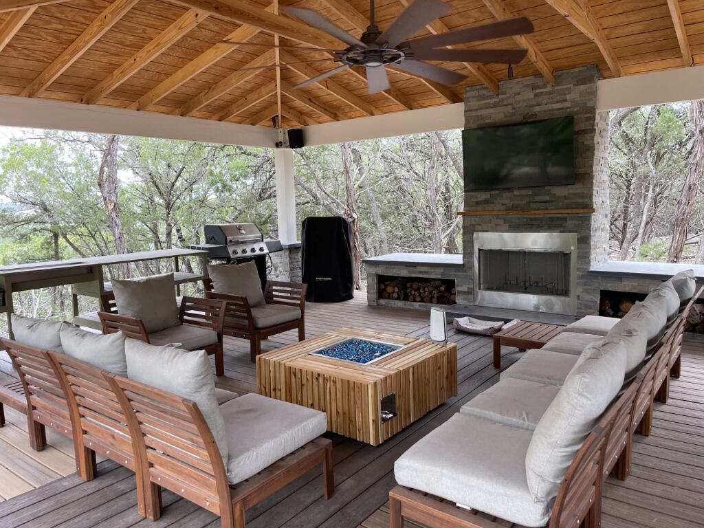 Covered patio with wood ceiling, cozy seating around a fire pit, stone fireplace and TV, barbecue grill, and lush trees.