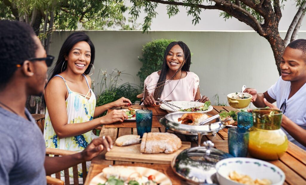 Four people enjoy a meal at a wooden table on an outdoor patio, surrounded by trees and greenery, sharing bread and salad with smiles.