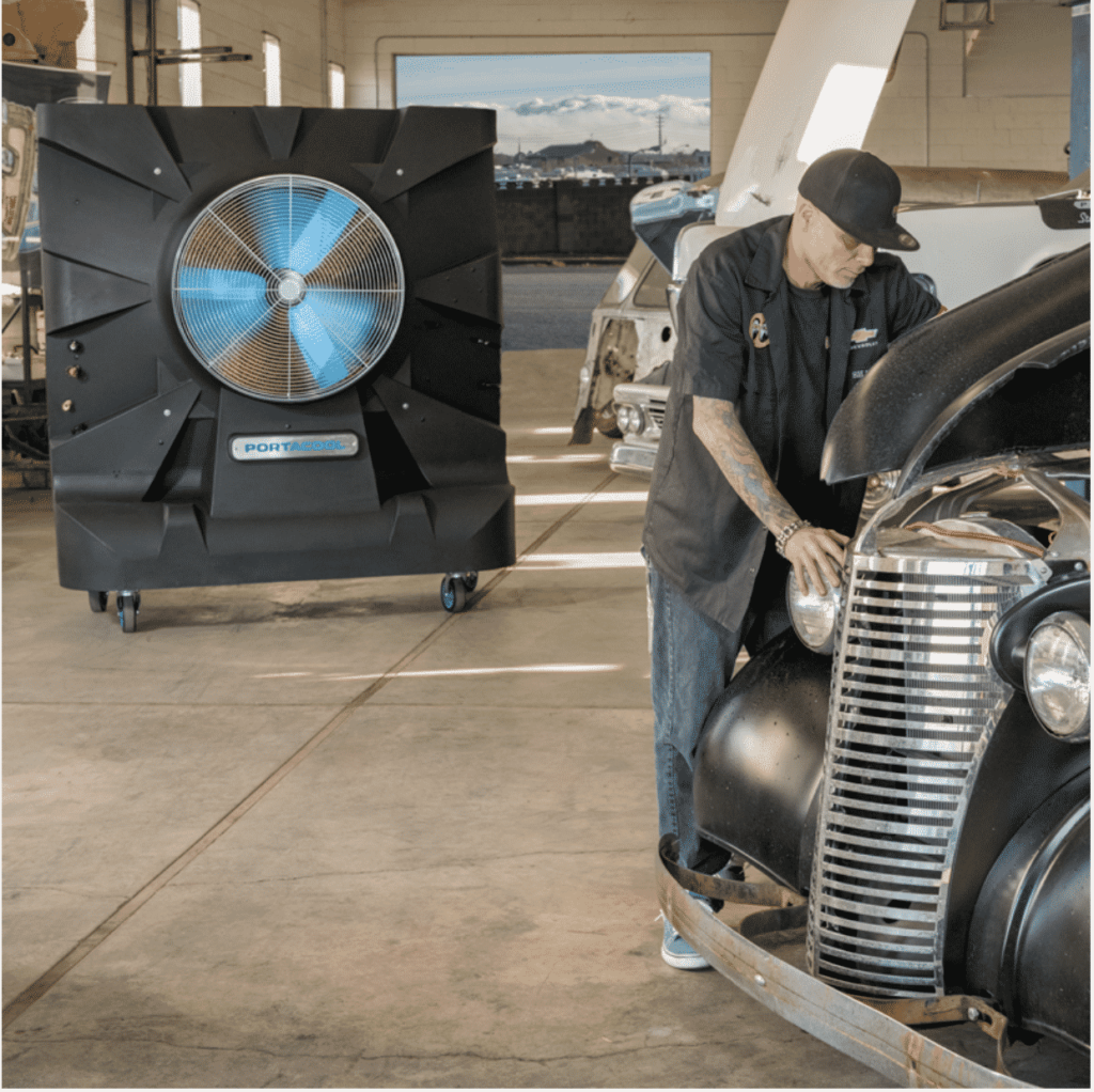 A person in a cap and dark shirt cleans a classic car’s headlight. A Hurricane with blue blades spins nearby, as sunlight streams through the open garage door.