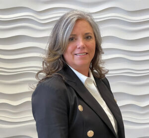 Jennifer Wyatt, a woman with long gray hair, smiles in a black blazer and white shirt against a textured wavy-patterned wall.