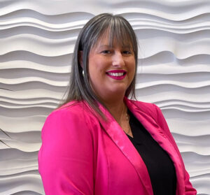 Jennifer Wyatt, with long gray hair, wears a bright pink blazer and black top while smiling warmly against a textured white wall with wavy patterns.