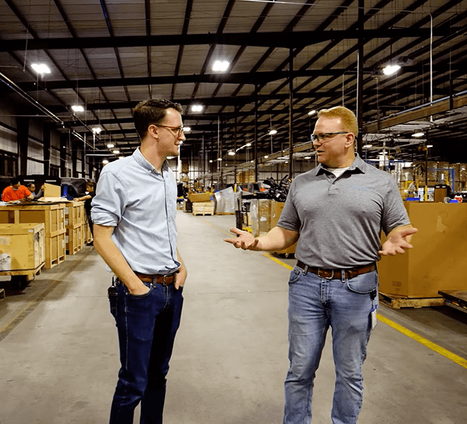 In a warehouse, two men in casual shirts and jeans chat under industrial lighting near a stack of "Made in America" Portacool units.