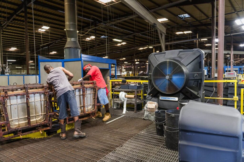 Two workers handle large metal containers in a factory, dressed casually. An Apex hums nearby in the industrial setting with exposed beams and equipment.