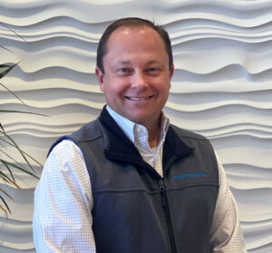A smiling man in a gray logo vest stands against a wavy white wall, with short hair and a checkered shirt. Plant leaves are visible to his left.
