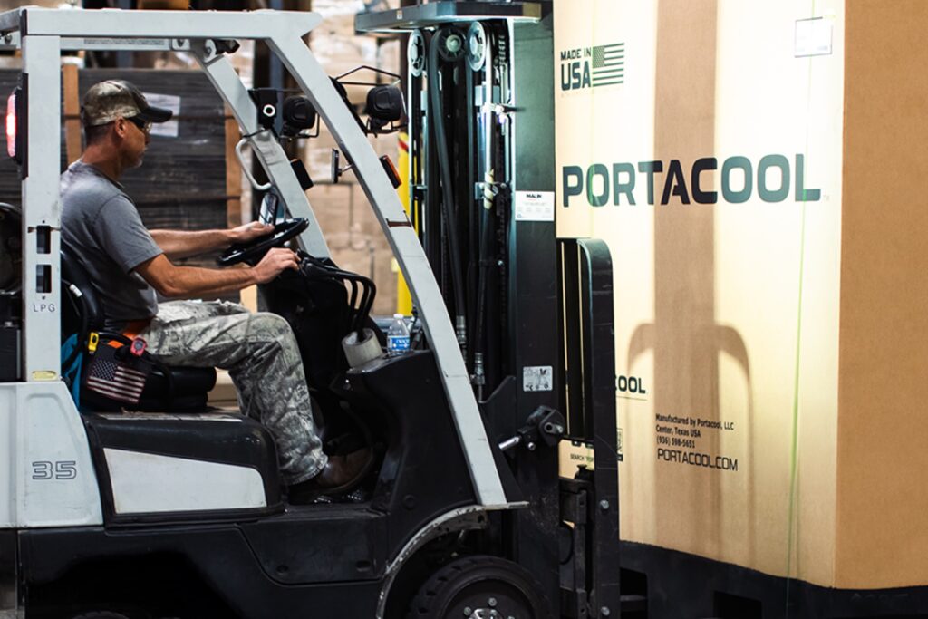 A person in camo, cap, and sunglasses operates a forklift moving a "Portacool" box in a warehouse.