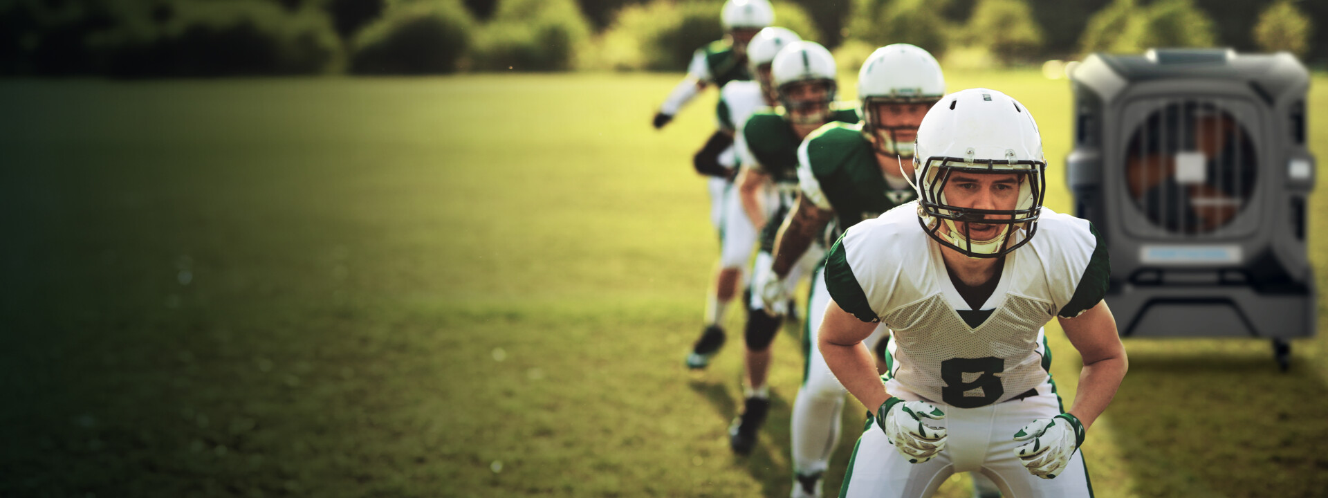 Football players in white and green uniforms prepare on the field. Player 6 leads, helmet on. A large metallic crate sits on the grass in the background.