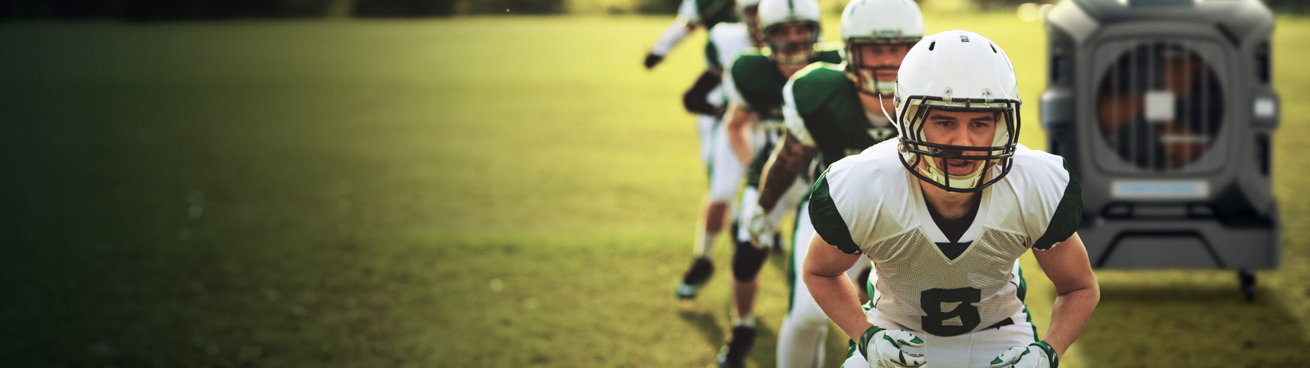 Football players in white and green uniforms prepare on the field. Player 6 leads, helmet on. A large metallic crate sits on the grass in the background.